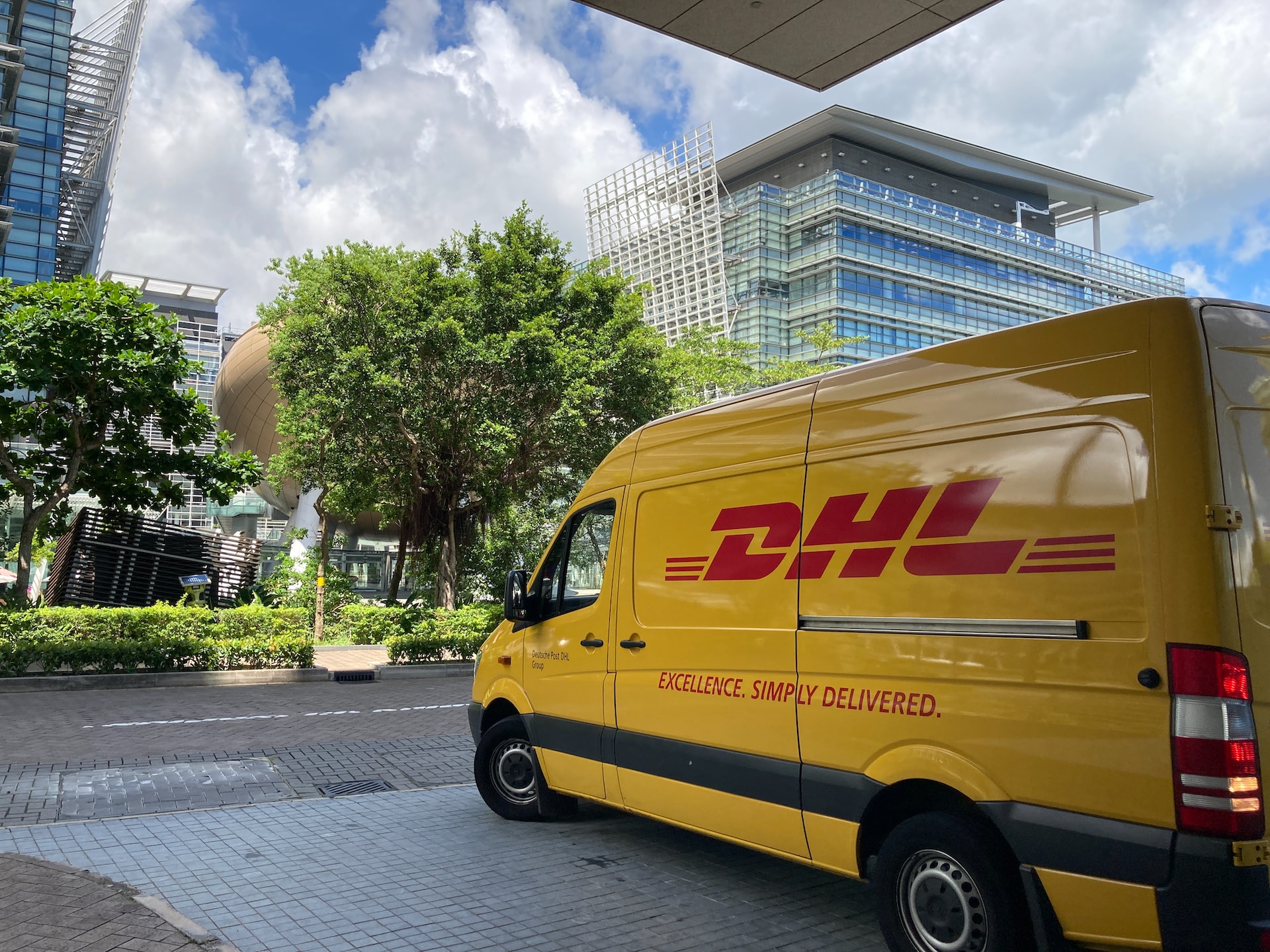 A yellow DHL van pulls out onto a street, there are high rise buildings in the background