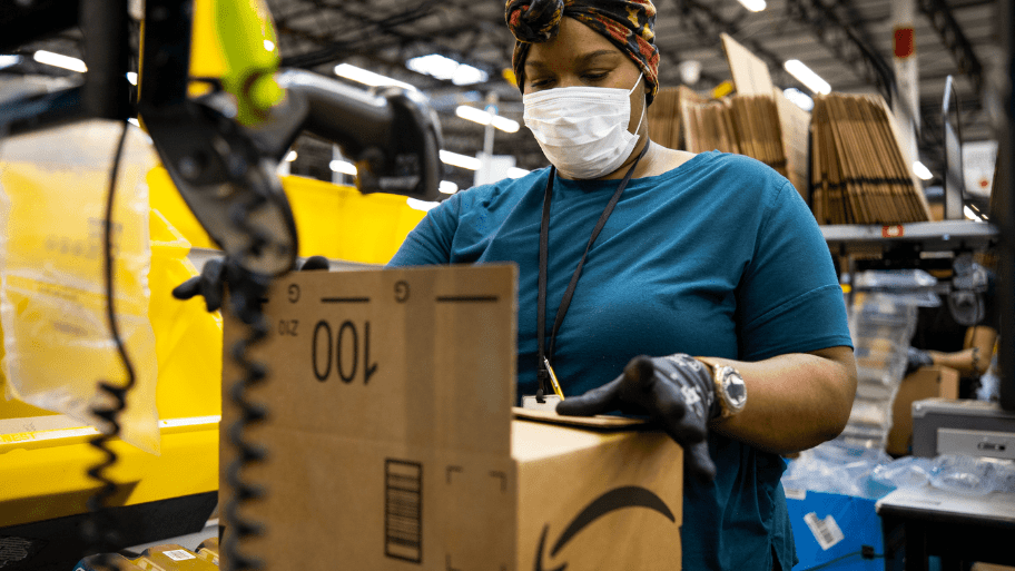 An Amazon worker stands in an Amazon warehouse packing an order into a box, The worker is wearing a blue shirt, white face mask, and a colorful head covering. She also wears gloves.