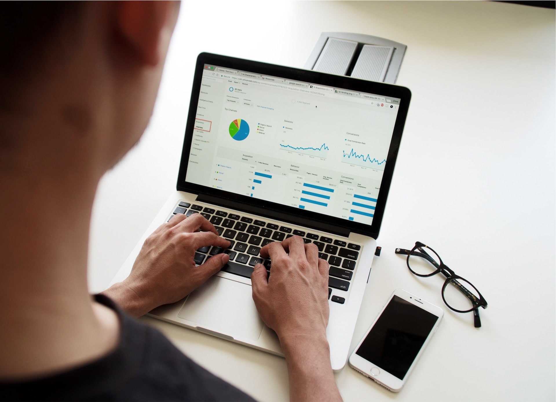 An over-shoulder shot of a man using a laptop, the screen shows Google Analytics for a website. Next to the computer as a set of reading glasses and a smartphone