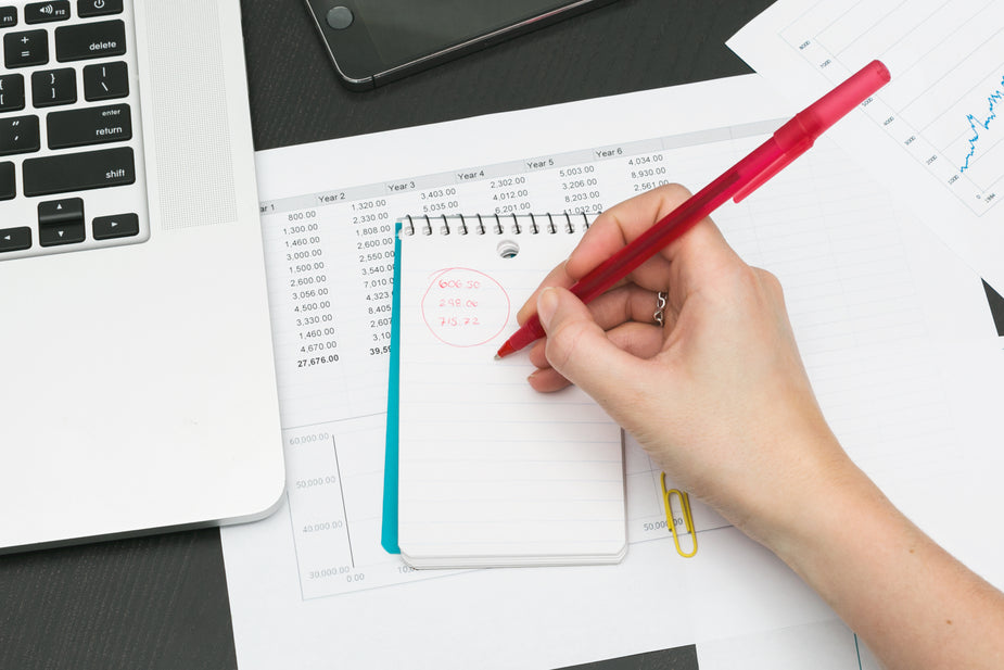 A hand holds a pen over a notepad doing budgeting, there are papers with spreadsheets and an open laptop nearby