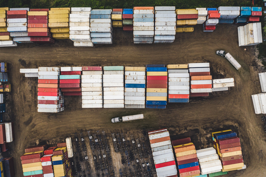 A overhead shot of colorful shipping containers stacked in rows. Two trucks drive on a dirt road between the rows.