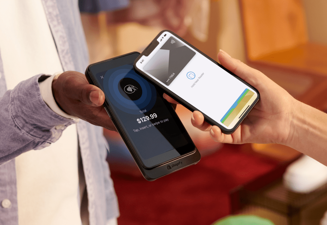 one hand holds a Shopify POS Go device indicating the total to be paid is $129.99. Another hand holds a smart phone over the device with their card open on the screen, paying for an item via Tap to Pay