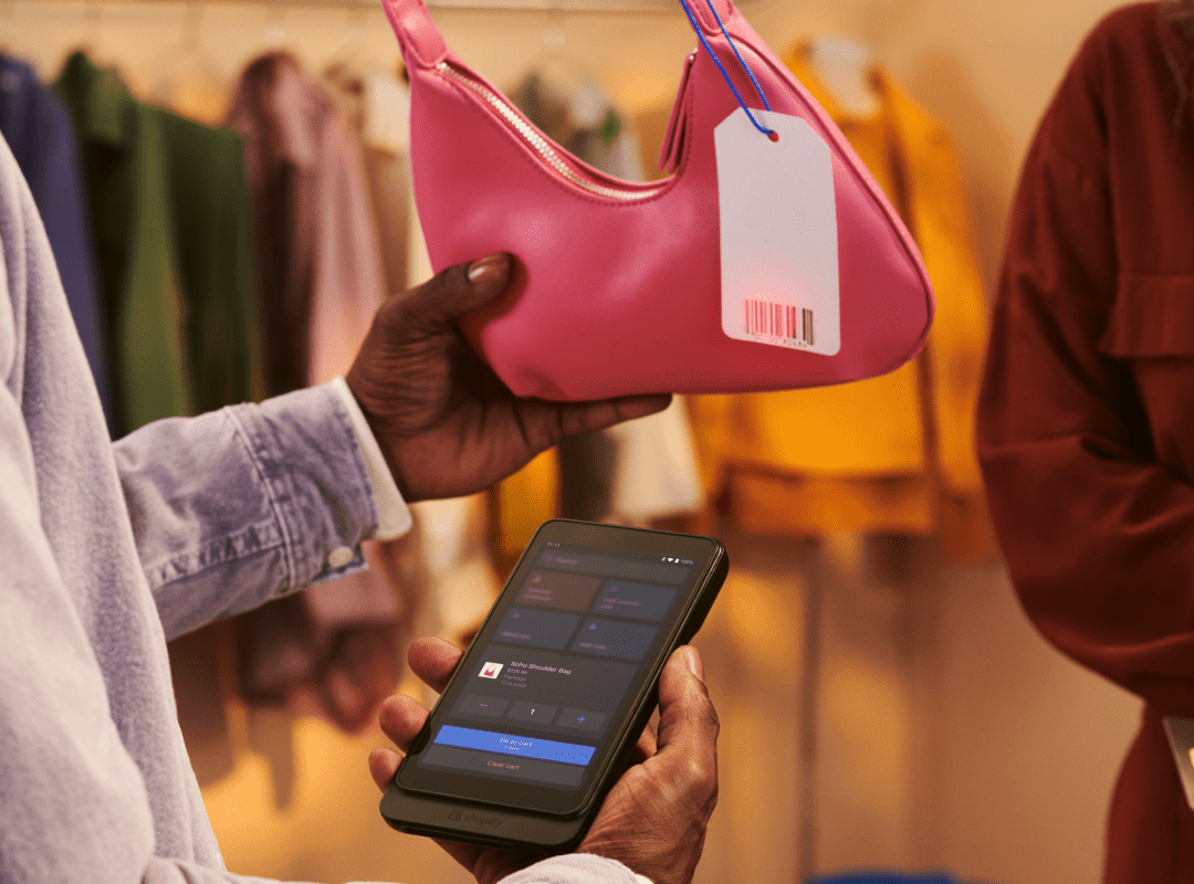 one hand holds a Shopify POS Go device and scans the barcode of a pink handbag