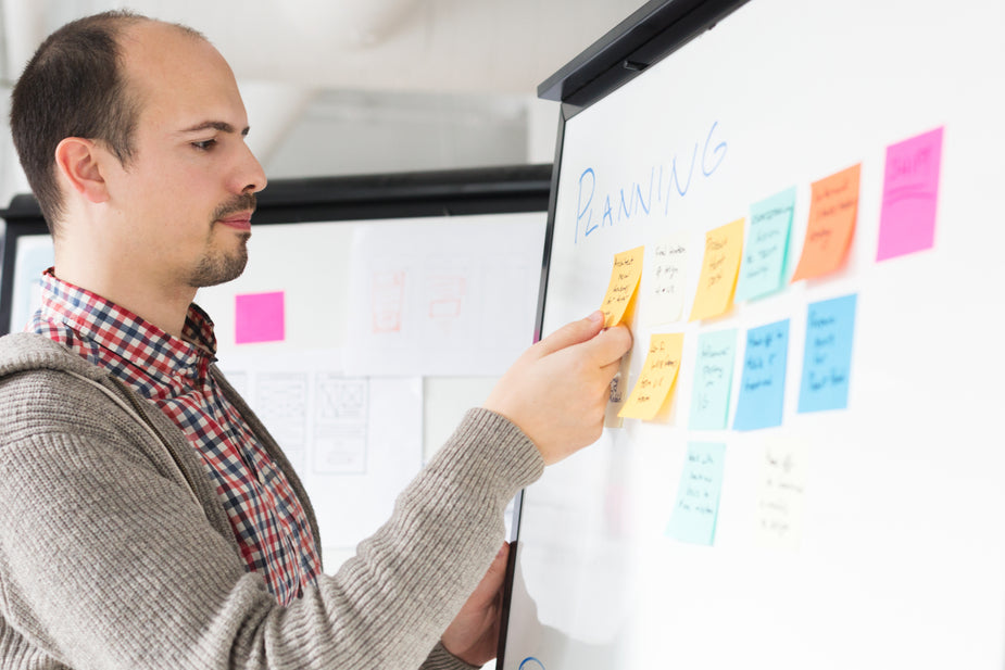 A balding man with a goatee stands in front of a whiteboard with the word “PLANNING” written on it and several colorful Post-It notes. The man is moving a Post-It note.
