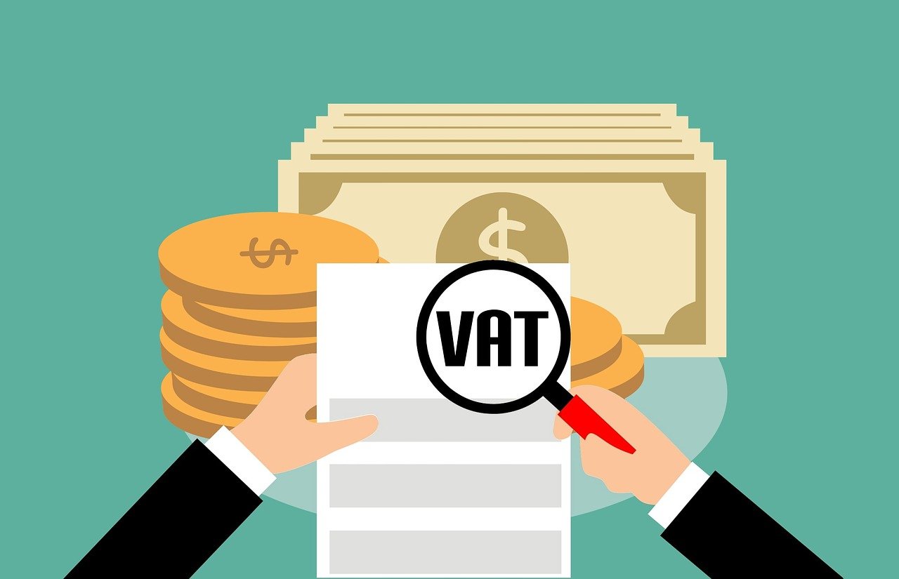 An illustration indicating VAT A cartoon illustration of a hand holding a magnifying glass over a piece of paper. The magnifying glass enlarges the letters 'VAT'. There are bank notes and coins in the background