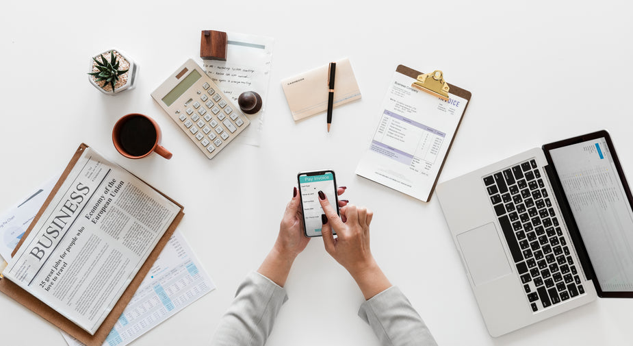 an overhead shot of women’s hands holding a phone, tapping the screen. On the desk behind the phone is a calculator, clipboard, open computer, and business newspaper. It look like they are sorting out their accounting.