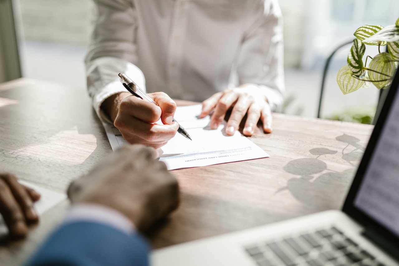 A shot of a woman’s hands signing a paper document. In the foreground you can see out of focus objects including a laptop and a man’s hands